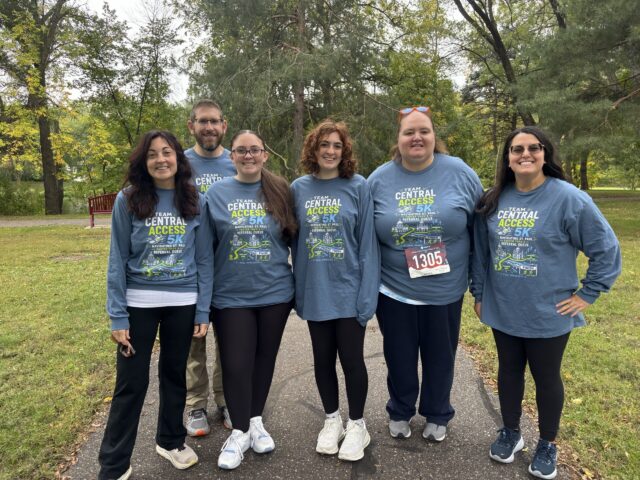 Six adults in teal Team Central Access shirts posing together on a park path with trees in the background.